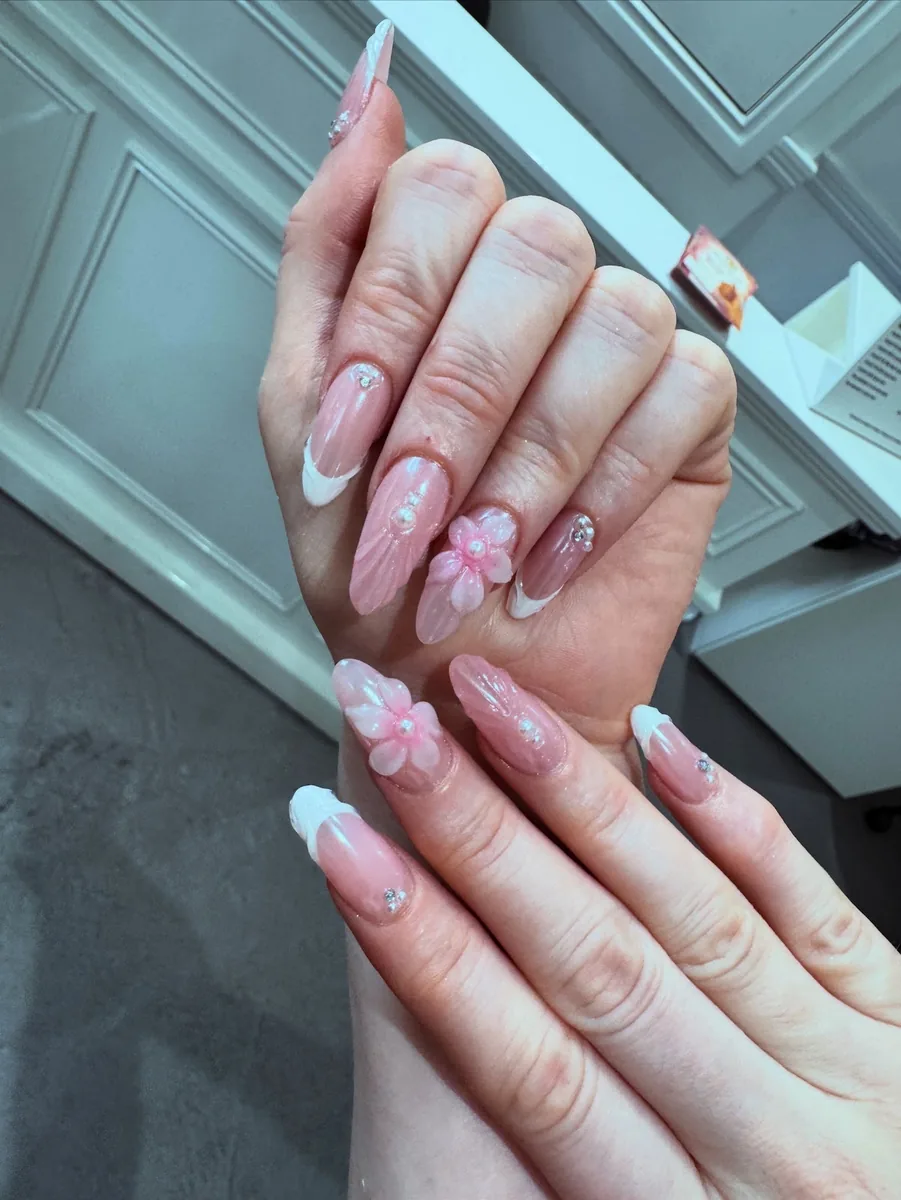 A finished set of long pink and white ombre nails with 3D floral art, held up against a sage green panelled wall.