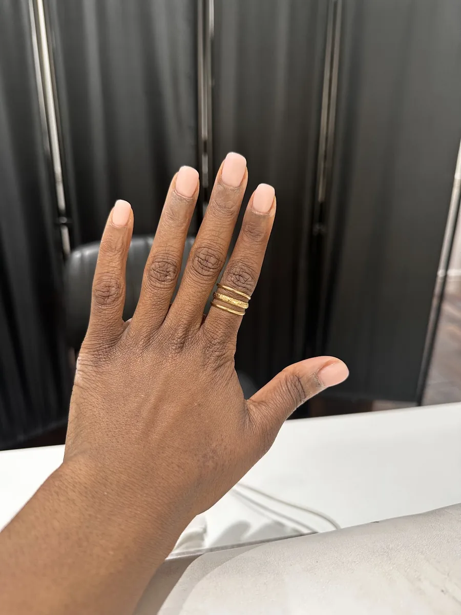 An open hand showing a completed short natural nail set, worn with gold rings.