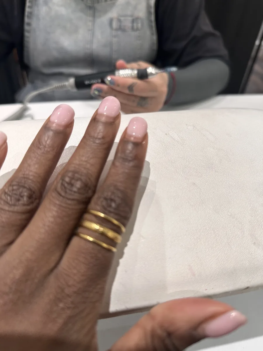 A hand with a soft pink manicure and stacked gold rings resting on the salon table.