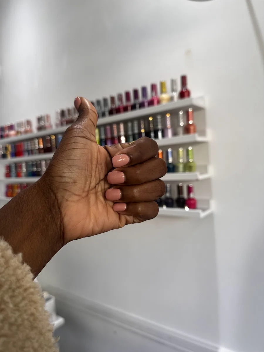 A closed hand with short dark glossy nails held in front of the salon's rack of colour bottles.